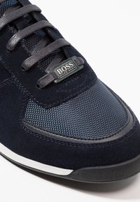 Marinblå sneakers med en blandning av mocka och mesh-texturer, med mörka snören och en silverfärgad logotyp. Inslag inkluderar en vit sula.