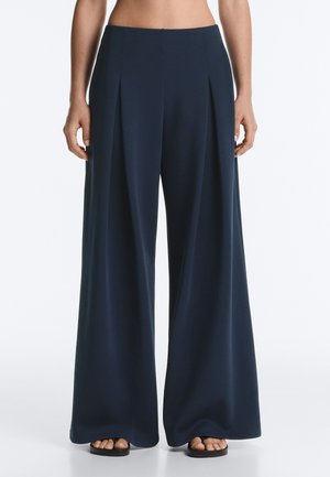 Femme portant un pantalon taille haute bleu marine à jambes larges avec des plis sur le devant et des sandales noires à bout ouvert, debout sur un fond blanc.