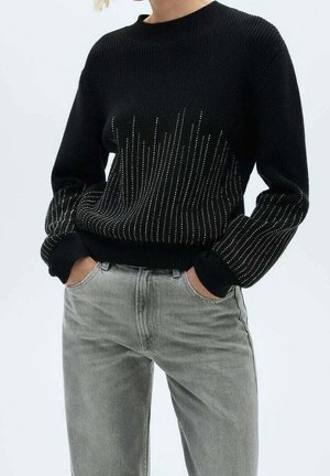 Schwarzer Strickpullover mit weißem gepunktetem vertikalem Linienmuster von der Taille bis zur Brust und an den Ärmeln, kombiniert mit hellgrauen Jeans.