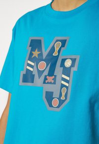 Camiseta de algodón turquesa con un gran gráfico de las letras "MJ" en azul marino, con varios iconos deportivos y rayas en color crema.