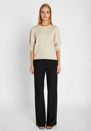 Ung kvinde står oprejst iført en beige kortærmet sweater, sorte bukser med vide ben og sorte åbne tåhæle mod en ensfarvet baggrund.