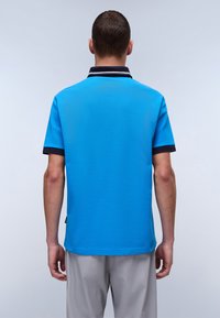 Pikétröja i ljusblått tyg med marinblå krage och ärmslut. Kort ärm och rak nederkant, med en liten sidologotyp detalj.