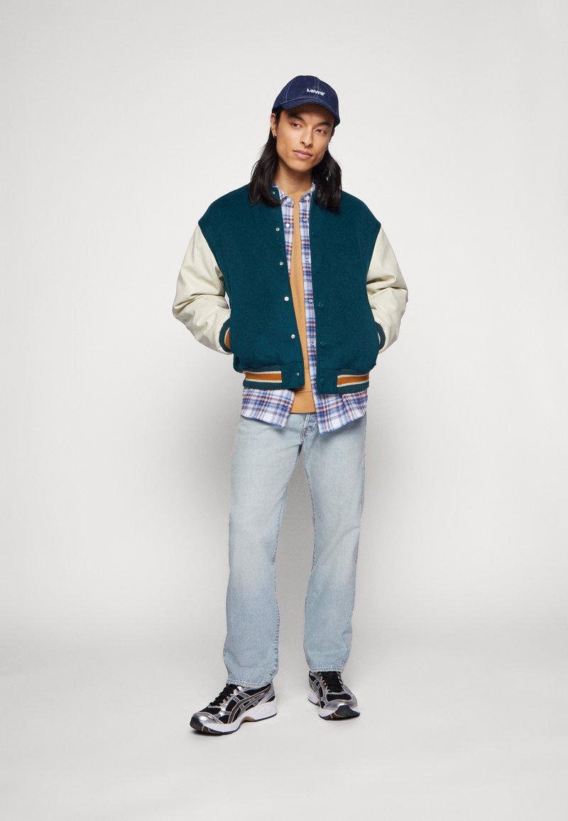 Teal- und cremefarbener College-Jacke über einem karierten Hemd, kombiniert mit hellen Jeans und metallischen Laufschuhen. Eine marineblaue Kappe mit Branding rundet den Look ab.