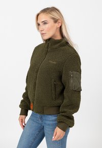 Zielona oliwkowa bluza polarowa z wysokim kołnierzem, zapinaną na zamek kieszenią na lewym rękawie oraz ściągaczami na rękawach. Posiada teksturowaną powierzchnię.
