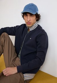 Navy zip-up sweatshirt with ribbed cuffs and a checkered collar, paired with loose tan pants and a blue denim cap. Simple, modern design.