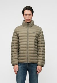 Tamsiai žalia pūkinė striukė su aukštu apykakle, horizontaliu siūlių raštu, priekiniais užtrauktukais ir subtiliu prekių ženklo ženklu. Sukomplektuota su mėlynomis džinsinėmis kelnėmis.