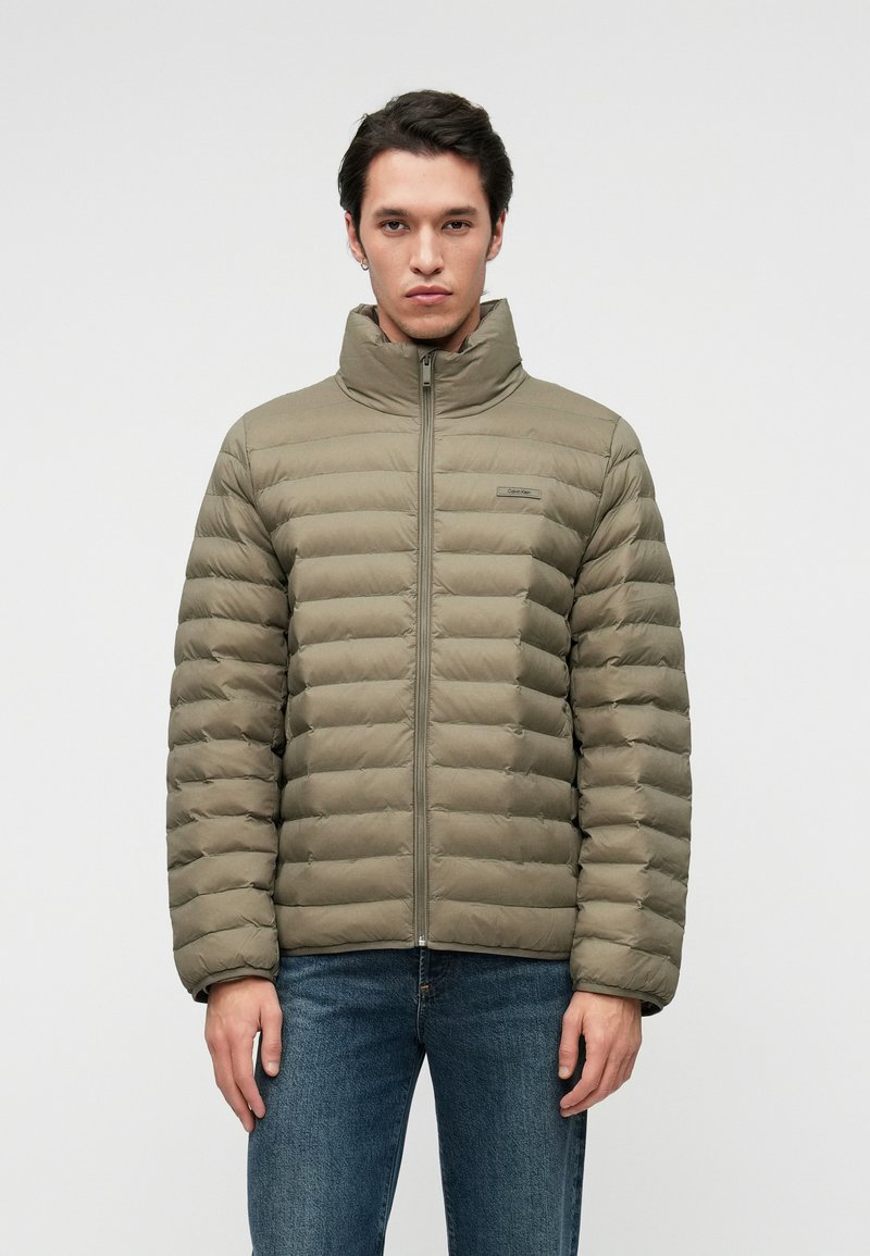 Tamsiai žalia pūkinė striukė su aukštu apykakle, horizontaliu siūlių raštu, priekiniais užtrauktukais ir subtiliu prekių ženklo ženklu. Sukomplektuota su mėlynomis džinsinėmis kelnėmis.