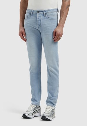Man die lichtblauwe slim-fit jeans en zilveren hardloopschoenen draagt, staand tegen een effen lichte achtergrond.