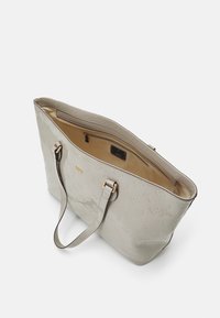 Sac fourre-tout beige avec motif floral embossé, ferrures dorées et doubles poignées. Le dessus ouvert révèle un intérieur marron clair avec une poche zippée.