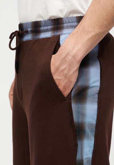 Άτομο που φοράει καφέ παντελόνια με μπλε καρό πλαϊνά πάνελ και κορδόνι στη μέση, το χέρι ακουμπά στην μπροστινή τσέπη.