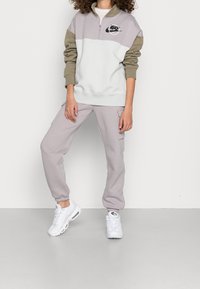 Lichtgrijs en taupekleurig trainingspak met een kwart-ritssluiting op de trui en cargobroek, voorzien van een zwart logo en elastische manchetten. Witte sneakers maken de look compleet.