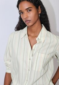 Gaiši zaļa, svītraina pogas aizdares krekls ar pasteļtoņu līnijām. Tam ir apkakle, priekšējā kabata un uzsietas piedurknes. Mīksta auduma tekstūra.