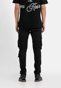 Zwarte cargobroek met meerdere zakken, nauwsluitende pasvorm en zachte stof. G gedragen met een zwart T-shirt en zwarte sneakers.