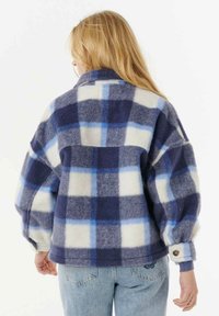 Vrouw met blond haar draagt een oversized blauwe en witte geruite fleecejas en lichtblauwe jeans, gezien vanachter.