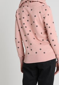 Felpa rosa con pois grigi, dotata di cappuccio, polsini e orlo a coste. Il tessuto appare morbido con una vestibilità casual e rilassata.