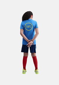 Maillot de sport bleu avec le logo "THFC" au dos, short bleu marine, chaussettes rouges montantes et chaussures de sport vert vif. Cheveux longs et bouclés.
