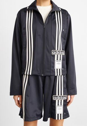 Persoon draagt een zwart Adidas-zip-up jack en bijpassende shorts met witte verticale strepen en logopatches aan de linkerzijde.