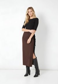 Top negro de manga tres cuartos combinado con una falda marrón de satén con una abertura lateral y detalle de flecos negros, complementada con botas negras hasta la rodilla.