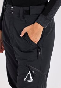 Schwarze Softshell-Hosen mit einer Vordertasche, seitlicher Reißverschlusstasche, doppelter Knopfverschluss und einem Klettverschluss am Bund.
