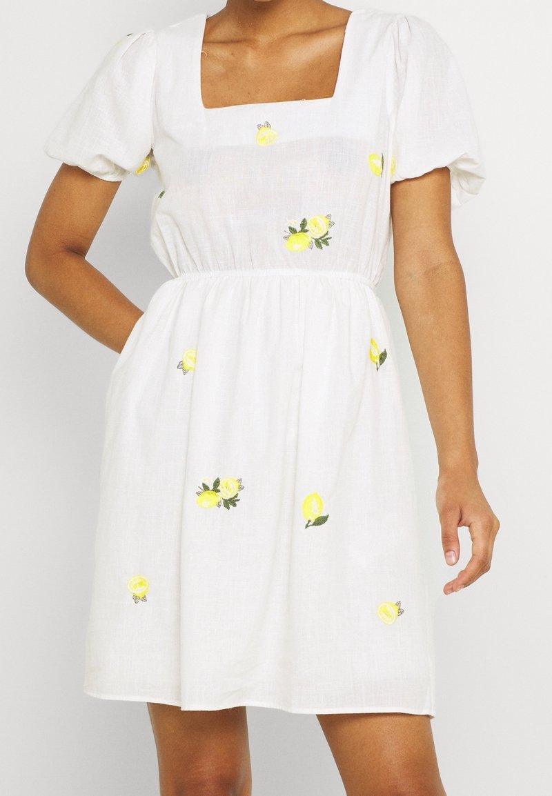 Weißes Kleid mit quadratischem Ausschnitt, kurzen Puffärmeln und bestickten gelben Zitronen und grünen Blättern. Leichter Stoff mit geraffter Taille.