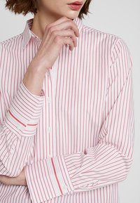 Chemise blanche boutonnée avec fines rayures rouges, dotée d'un col classique, de manches longues avec poignets à boutons, et d'une texture de tissu lisse.