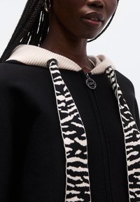 Sudadera negra con cremallera, capucha acanalada en beige, cordones llamativos en blanco y negro y cierre con cremallera elegante con tirador con logo.