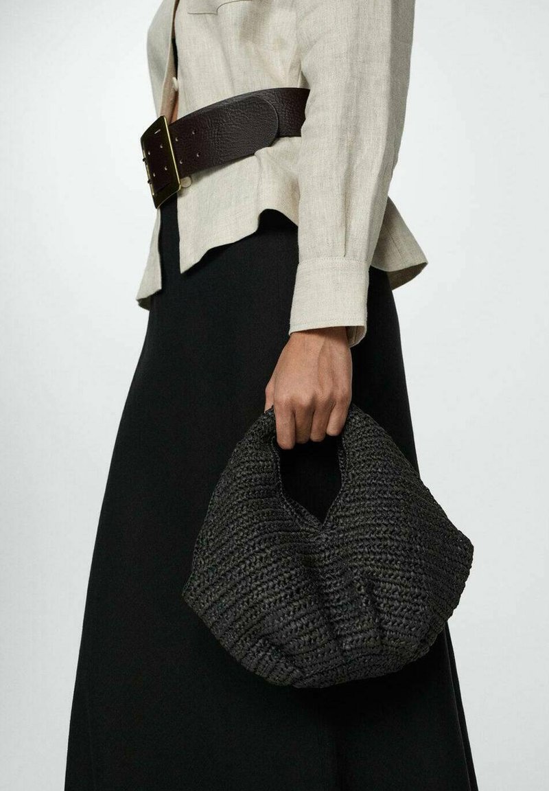 Schwarze gewebte Tasche mit strukturiertem Finish und einzigartiger Form, die von einer Hand gehalten wird. Die Tasche hat einen runden oberen Teil und einen kleinen Henkel.