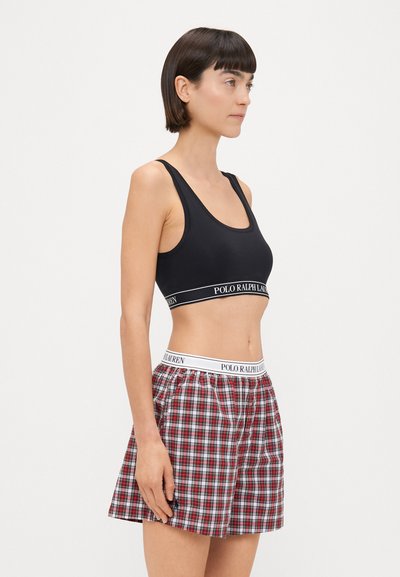 Μαύρο αθλητικό σουτιέν με λευκή λωρίδα λογοτύπου, συνδυασμένο με κόκκινες και καρό βερμούδες που διαθέτουν λευκή ελαστική ζώνη με λογότυπο.