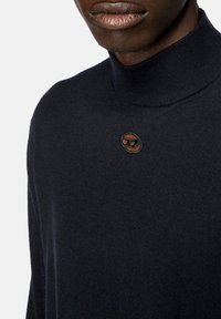 Maglione a collo alto di un blu navy scuro, realizzato in morbido tessuto, con colletto a coste e un piccolo logo circolare ricamato davanti.