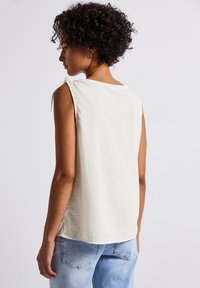 A woman with curly, short hair is seen from behind, wearing a light-colored sleeveless top and distressed blue jeans.
