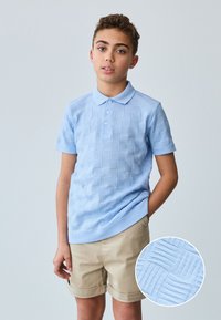 Vaaleansininen neulottu poolopaita, jossa on teksturoitu ruutukuvio, lyhyet hihat ja kauluksellinen pääntie. Asua täydentävät beige shortsit.