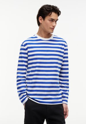 Νεαρός άνδρας που κοιτάζει δεξιά, φορώντας μπλε και άσπρη ριγέ μακρυμάνικη μπλούζα και μαύρο παντελόνι, με τα χέρια στις τσέπες.