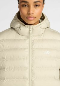 Chaqueta de pluma color beige claro con cremallera en la parte delantera, capucha y diseño de costuras horizontales. Presenta un logotipo blanco de New Balance en el lado izquierdo del pecho.