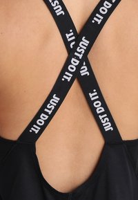 Haut de sport noir avec des bretelles croisées portant le texte « JUST DO IT » en blanc. Le design du dos est minimaliste avec une texture lisse.