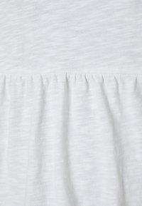 Tecido branco texturizado com bainha em babados, apresentando um padrão sutil e irregular. Material suave e leve visível em vista aproximada.