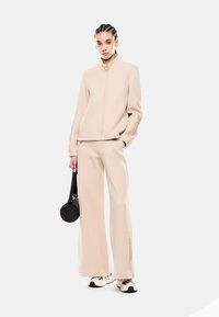 Chaqueta beige con cremallera y cuello alto, combinada con pantalones de pierna ancha. Detalle de marca sutil en los pantalones, complementado con un bolso negro redondo y zapatillas.