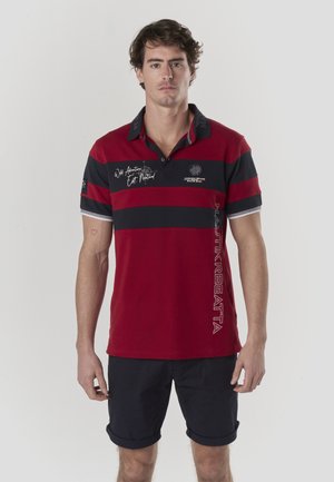 Polo de rayas rojas y azul marino con mangas cortas, cuello y detalles de texto. El material parece suave, diseñado para uso casual. Se ven pantalones cortos azul marino debajo.