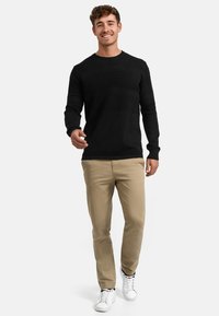 Musta, teksturoitu neulepaita yhdistettynä beigen värisiin housuihin ja valkoisiin lenkkareihin, jossa on istuva leikkaus ja pyöreä pääntie.