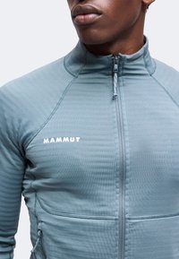 Gros plan d'une personne portant une veste Mammut bleu-vert zippée avec le logo de la marque visible sur la poitrine et un col montant.