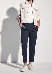 Helle beige, verkürzte Jacke mit Knopfleiste und Brusttaschen, kombiniert mit dunkelblauen Hosen und weißen Sportsneakern. Glatte Textur.