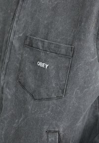 Dark gray denim shirt featuring a chest pocket with a white "OBEY" logo. The fabric has a textured finish with subtle fading.
