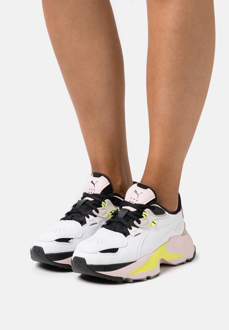 Białe i czarne sneakersy z różowymi akcentami, teksturowany materiał, gruba podeszwa i wyraźny żółty akcent po boku. Sznurówki z czarnymi elementami.
