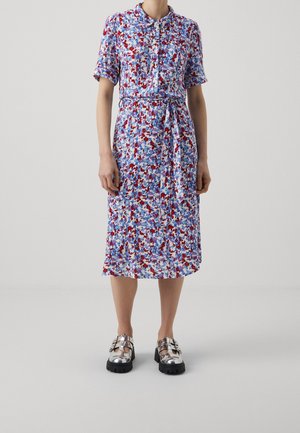 Femme portant une robe fleurie à manches courtes et longueur genou, avec une ceinture à la taille et des chaussures argentées métalliques à boucle, sur un fond uni.