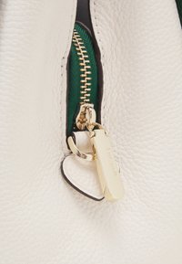 Biała skórzana torba z złotym zamkiem błyskawicznym z zielonym akcentem i złotą metką z napisem „kate spade”. Teksturowana powierzchnia i kontrastujące detale.