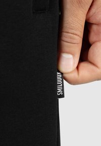 Prenda de tela negra con un primer plano de una mano sosteniendo una etiqueta. La etiqueta tiene impreso "SMLLTDXX". El material parece suave y texturizado.