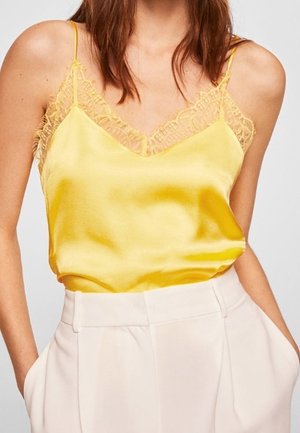 Vrouw in een heldere gele satijnen cami met kantafwerking en hoge witte gep tailored broek, met de handen in de zakken.