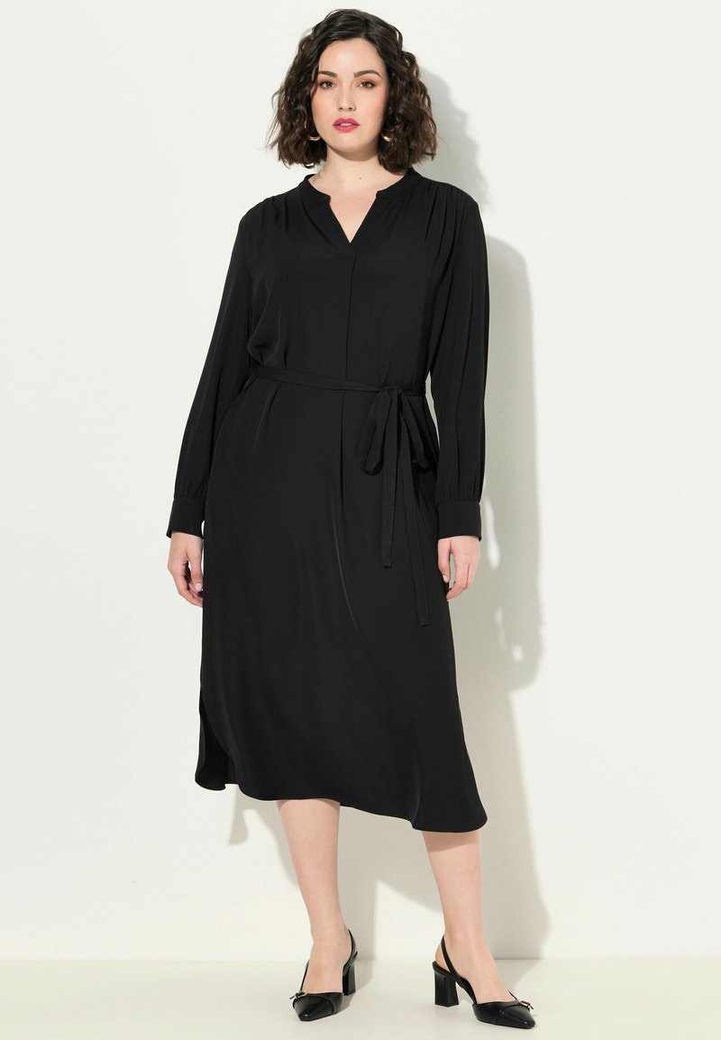 Vestido negro de manga larga con escote en V, cintura ceñida con un lazo, silueta fluida y aberturas laterales. Combinado con zapatos de tacón negros.