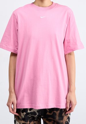 Personne portant un t-shirt Nike rose ample et un pantalon à imprimé léopard, debout devant un fond clair uni.