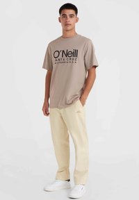 T-shirt en coton beige avec un grand imprimé noir "O'Neill SANTA CRUZ", associé à un pantalon jaune clair et des baskets blanches, debout contre un fond blanc.