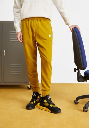 Osoba w mostkowych żółtych dresach Nike i czarno-żółtych butach Air Jordan stoi obok niebieskiego krzesła biurowego i metalowej szafki.